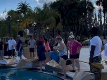 People are gathered by a poolside, preparing to use cardboard boats. Palm trees and a partly cloudy sky are in the background.