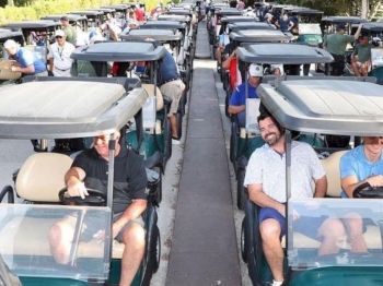 A large group of people is sitting in rows of golf carts on a path, likely preparing for a golf event, surrounded by greenery.