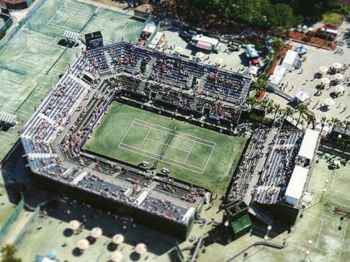 An aerial view of a sports complex featuring a central tennis court with surrounding stands and several outer courts, likely during an event.