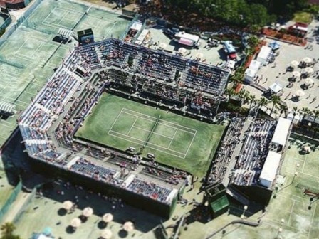 An aerial view of a sports complex featuring a central tennis court with surrounding stands and several outer courts, likely during an event.