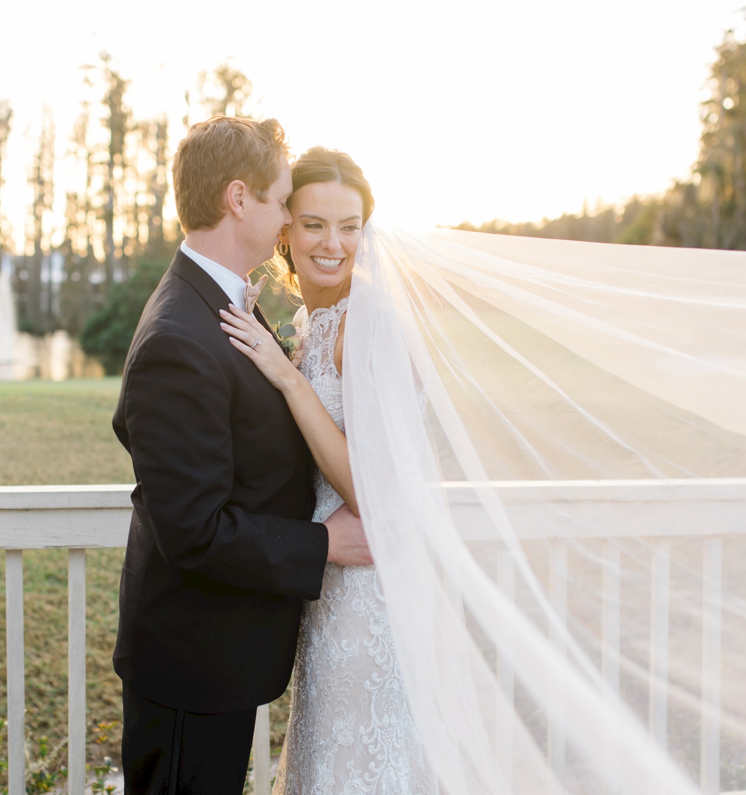 A couple stands on a bridge, embracing, with the bride's veil flowing in the breeze under a sunset backdrop.