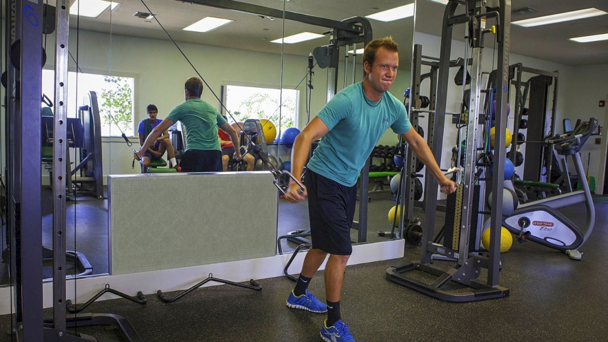 A person in a gym is using a cable machine for exercise while facing sideways. The background shows various gym equipment and a large mirror.