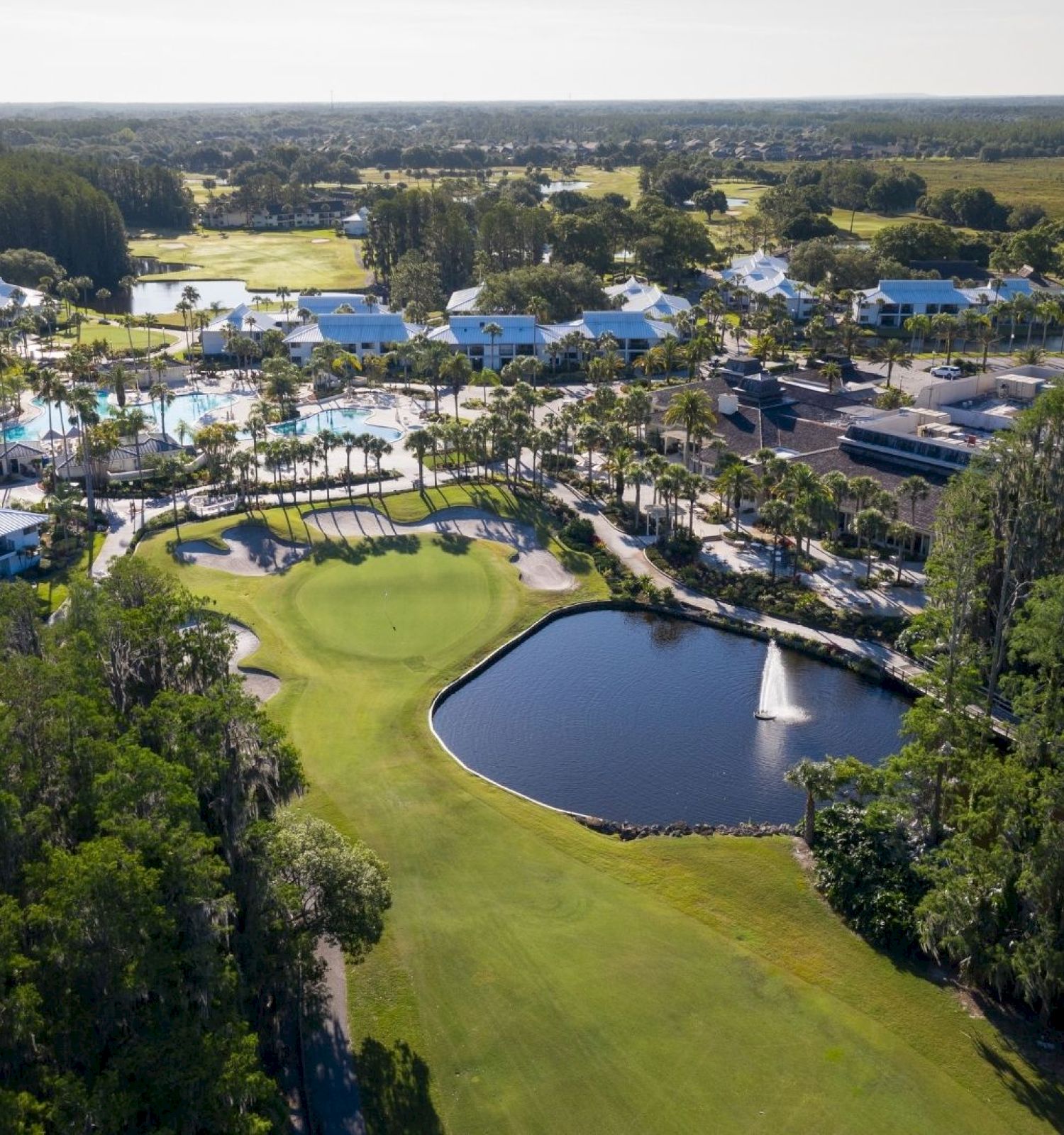 The image shows an aerial view of a golf course with a water feature, surrounded by buildings and greenery, extending to the horizon.