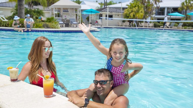 A woman is sitting by a pool with drinks, while a man and a child are enjoying the water; the child is on the man's shoulders, smiling and posing.