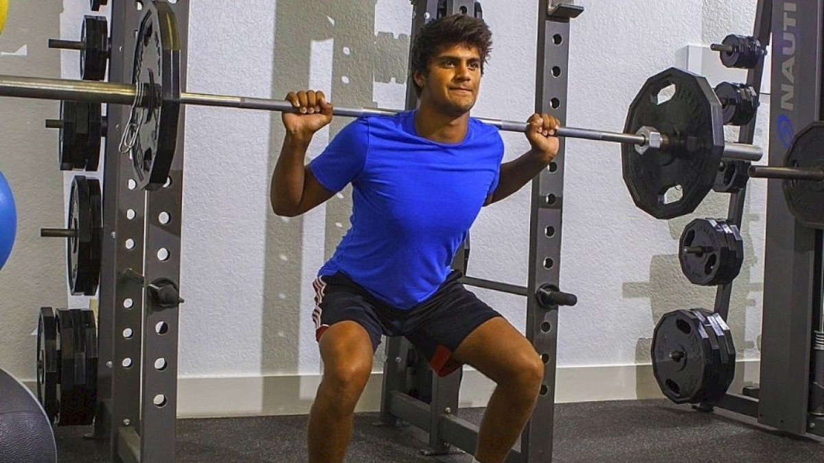 A person in a gym doing a barbell squat; they are wearing a blue shirt, black shorts, and orange shoes, using a squat rack for support during the exercise.