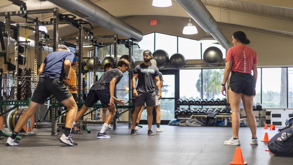 People are engaged in an indoor exercise session, with cones and gym equipment present in the background.