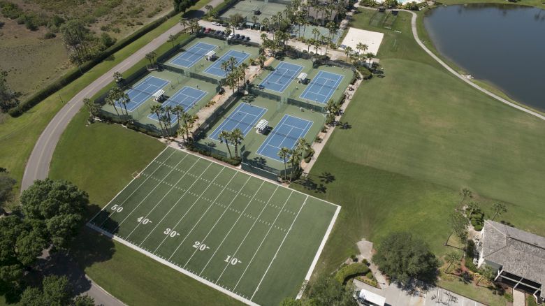 Aerial view of a sports complex with six tennis courts, a small football field, a lake, and nearby buildings under clear skies, ending the sentence.