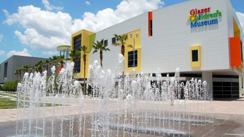 The image shows the exterior of Glazer Children's Museum with a splash fountain in the foreground, set against a bright, partly cloudy sky.