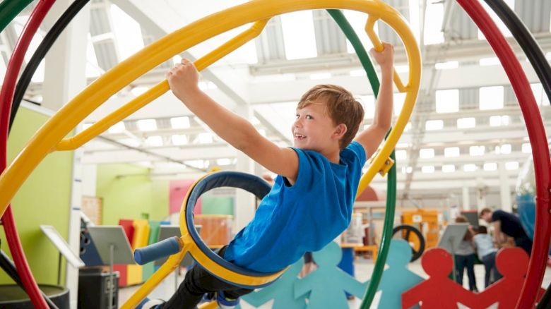 A smiling child is playing on a colorful climbing structure indoors, surrounded by large cutouts of people holding hands.