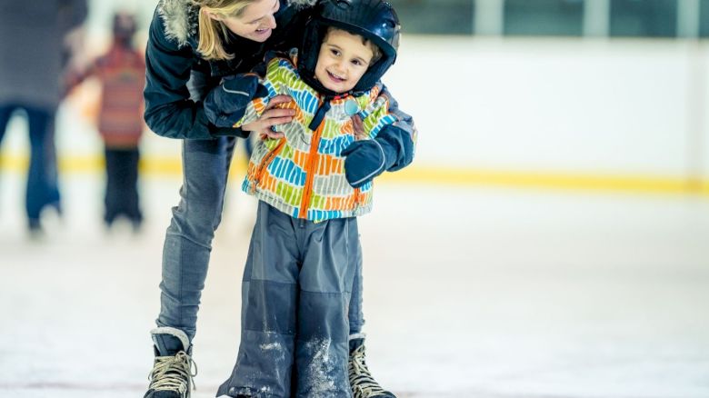 A woman helps a young child ice skate, both smiling, with the child wearing a helmet and colorful jacket on an indoor ice rink.
