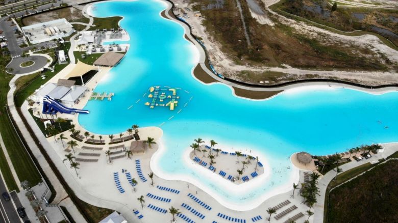 The image shows a large, lagoon-style swimming pool with water slides, floating inflatables, and numerous beach chairs. The area is surrounded by sandy beach.