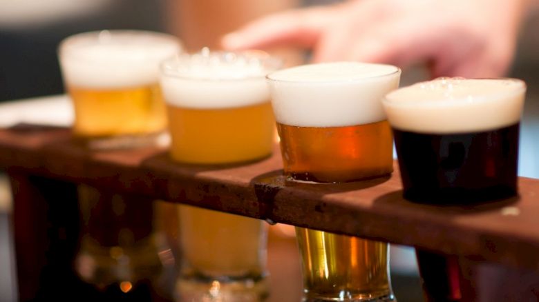 A wooden tray holds four different types of beer in small glasses, with varying colors and foam levels, while a hand reaches towards them.