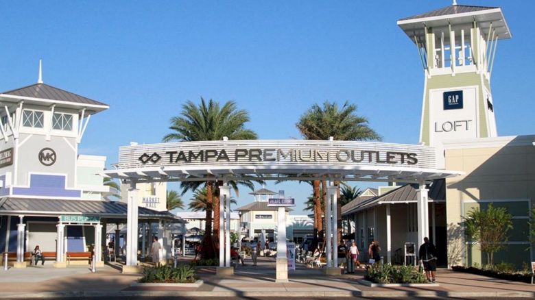 The image shows the entrance to Tampa Premium Outlets, featuring a sign, palm trees, and stores with the 'LOFT' building visible in the background.