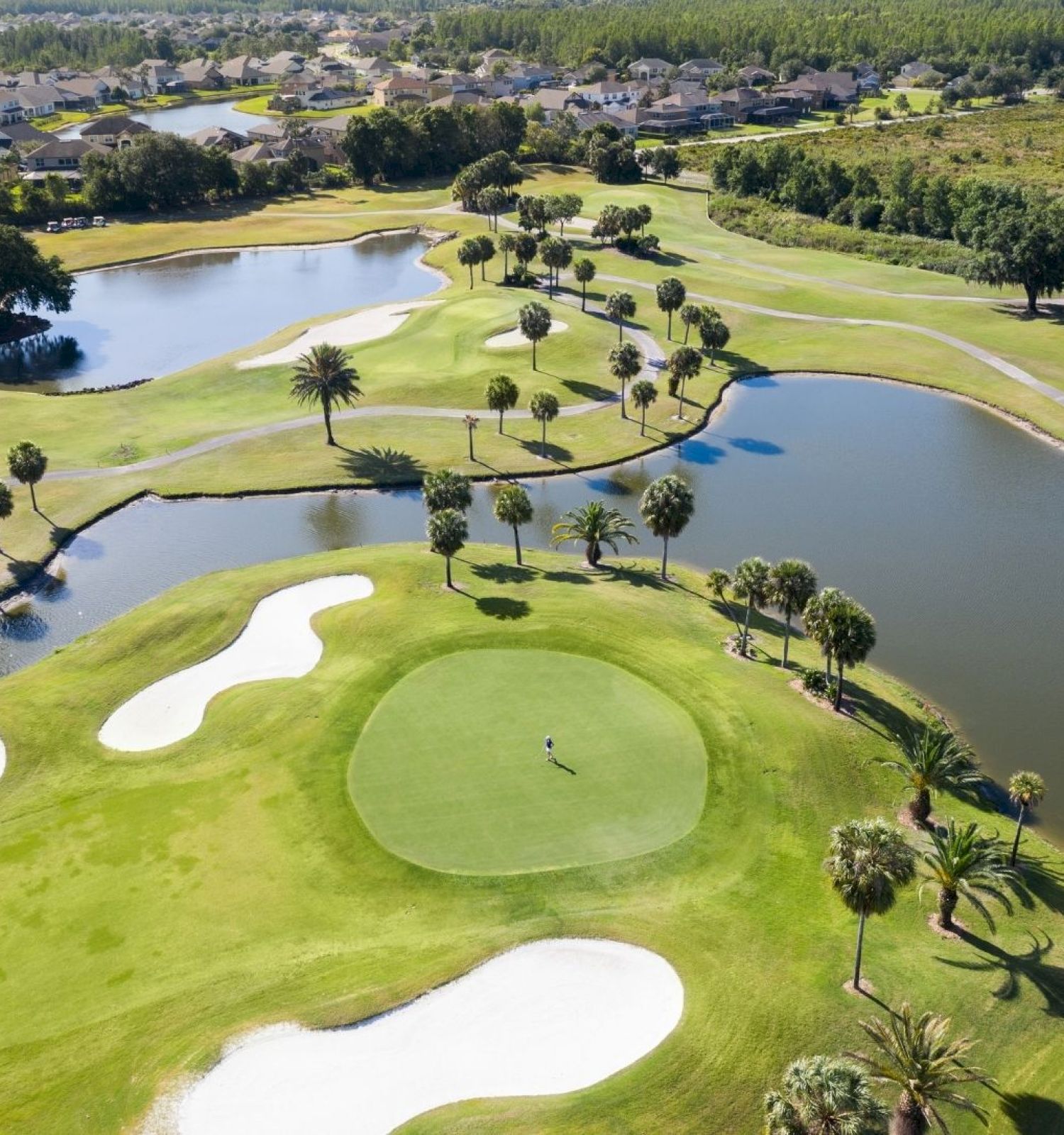 A scenic aerial view of a golf course featuring greens, sand bunkers, water hazards, palm trees, and surrounding residences in the background.