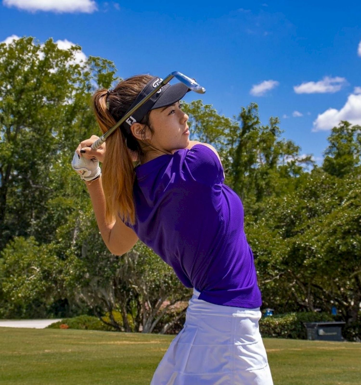 A person in a purple shirt and white skirt is swinging a golf club on a golf course under a blue sky with scattered clouds.