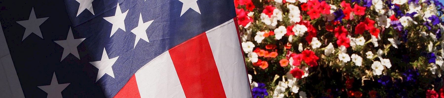 A United States flag is in the foreground, with a vibrant hanging basket of red, white, and purple flowers in the background against a clear blue sky.