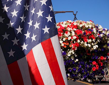 A United States flag is in the foreground, with a vibrant hanging basket of red, white, and purple flowers in the background against a clear blue sky.