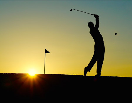 A silhouette of a golfer in mid-swing at sunset, with a flagstick in the background. The sun is low on the horizon, casting a warm glow.