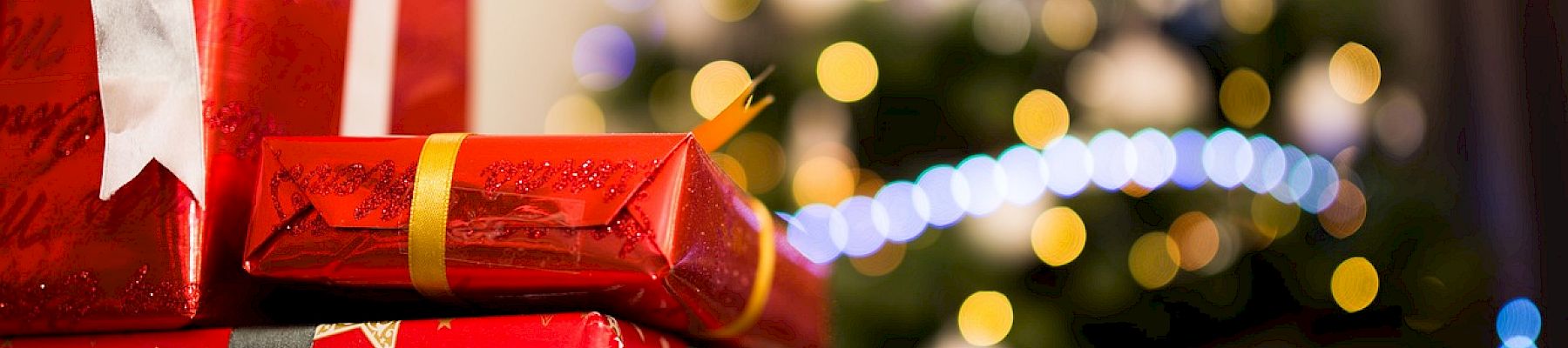 The image shows red and patterned gift boxes with ribbons in front of a decorated Christmas tree adorned with lights and ornaments.