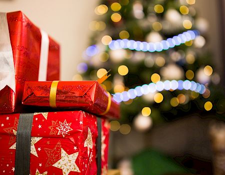 The image shows red and patterned gift boxes with ribbons in front of a decorated Christmas tree adorned with lights and ornaments.