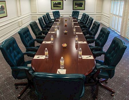The image shows a formal conference room with a long table, surrounded by twelve chairs, each set with a notepad and a water bottle.