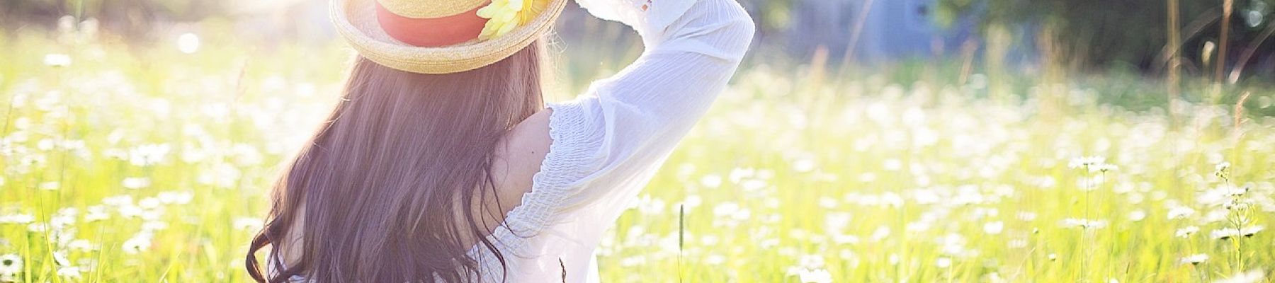 A person with long hair sits in a field of flowers, wearing a sunhat with a yellow flower, under a bright sunlit sky.