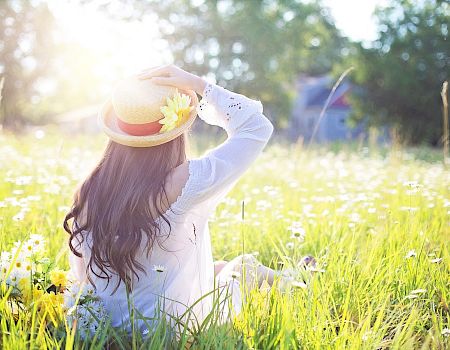 A person with long hair sits in a field of flowers, wearing a sunhat with a yellow flower, under a bright sunlit sky.