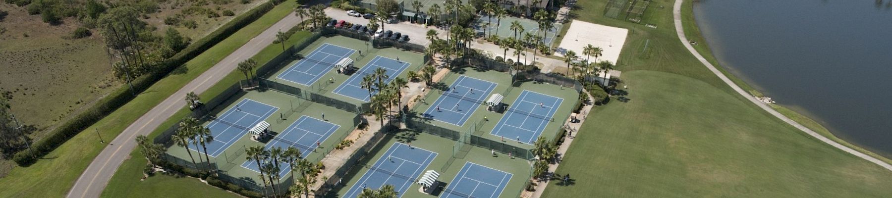 The image shows an aerial view of a sports complex with multiple tennis courts, a pool area, and adjacent green spaces near a water body, ending the sentence.