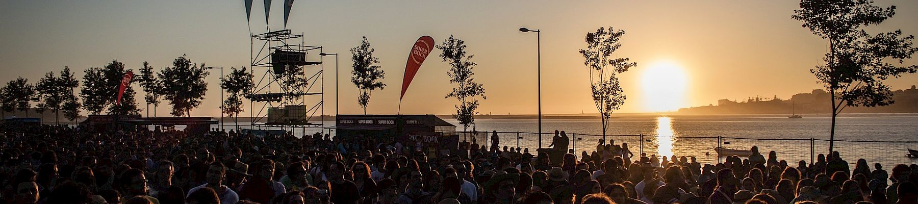 A large crowd gathers near a waterfront as the sun sets, with flags and a stage structure in the background, during an outdoor event.