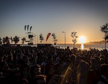 A large crowd gathers near a waterfront as the sun sets, with flags and a stage structure in the background, during an outdoor event.