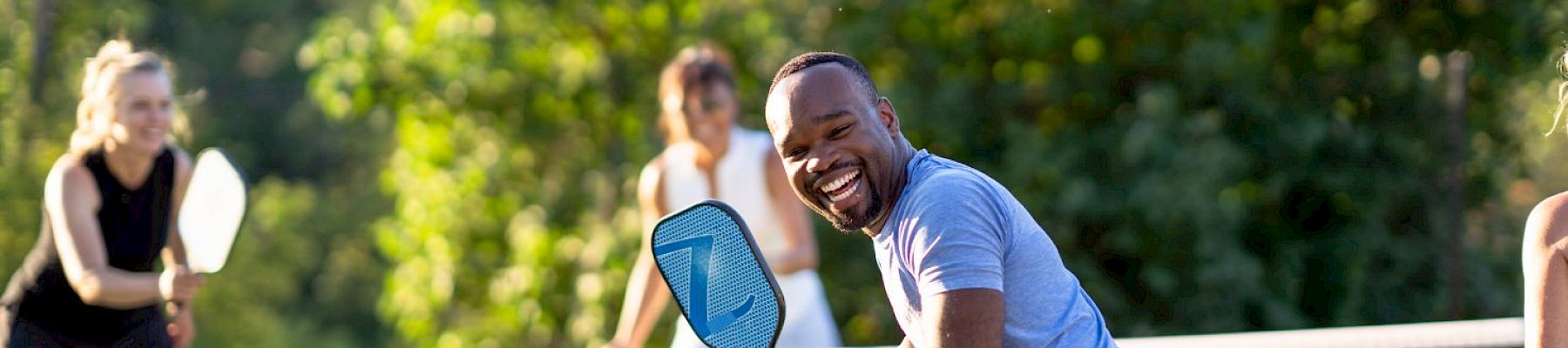 A group of people playing pickleball on an outdoor court, with three women and one man having fun and holding paddles, ending the sentence.