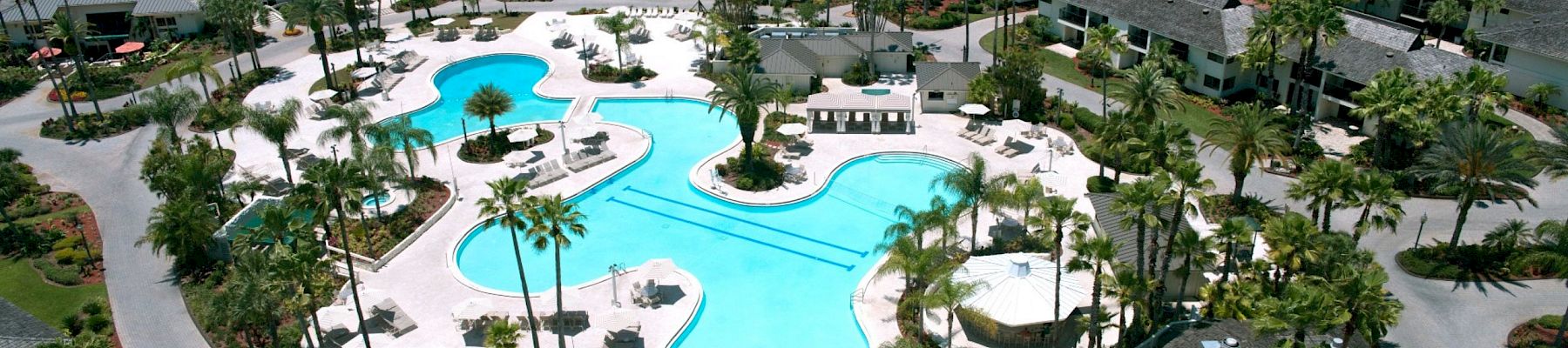 An aerial view of a resort featuring a large swimming pool, surrounded by lush greenery, palm trees, and various buildings.