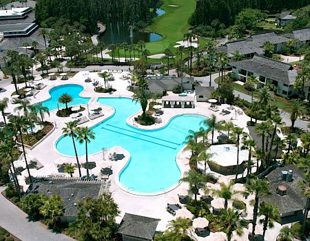 An aerial view of a resort featuring a large swimming pool, surrounded by lush greenery, palm trees, and various buildings.