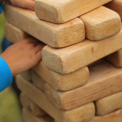 Several hands are playing with a large Jenga tower made of wooden blocks outdoors, focused on balancing the structure.