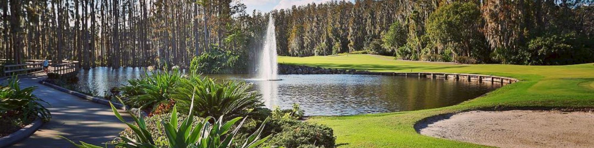 A serene golf course features a lake with a central fountain, surrounded by lush greenery, colorful plants, a bridge, and a sand trap.
