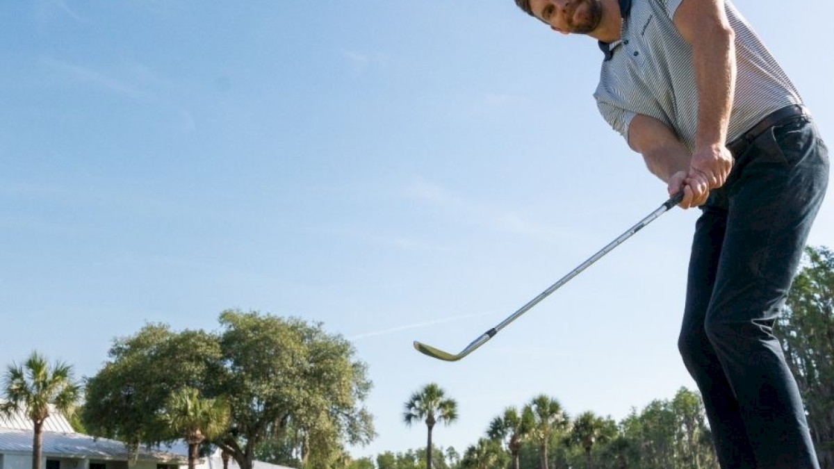 A man in a golf course swings a club at a ball, mid-air. Palm trees and a building are visible in the background under a clear blue sky.