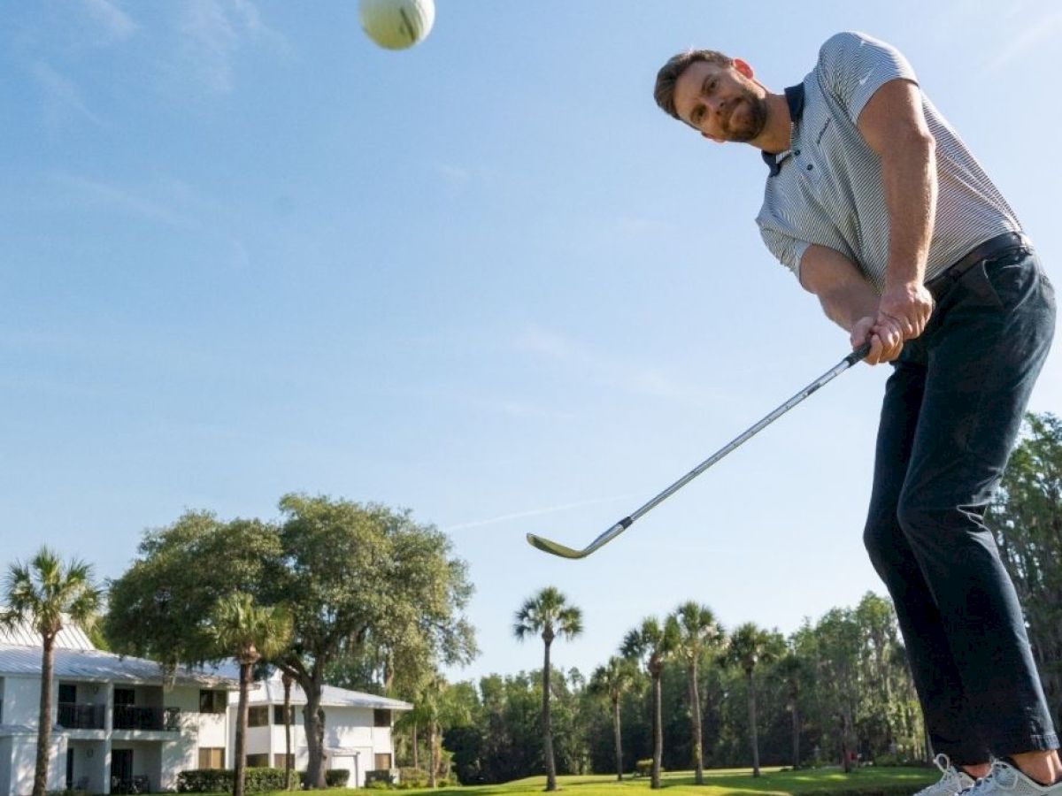 A man in a golf course swings a club at a ball, mid-air. Palm trees and a building are visible in the background under a clear blue sky.