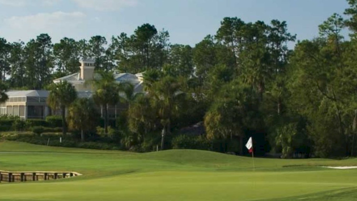 The image shows a golf course with a flag on the green, surrounded by trees and houses in the background under a clear sky.