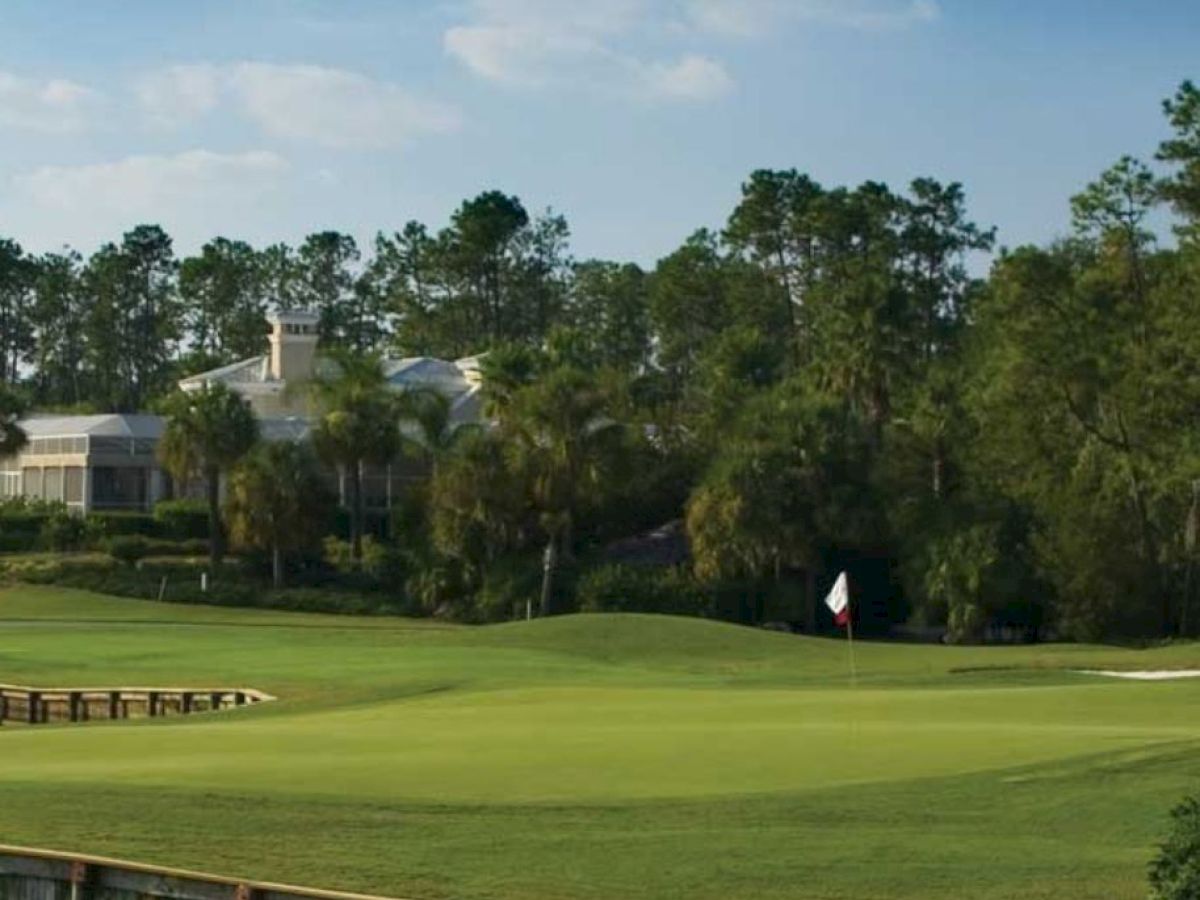 The image shows a golf course with a flag on the green, surrounded by trees and houses in the background under a clear sky.
