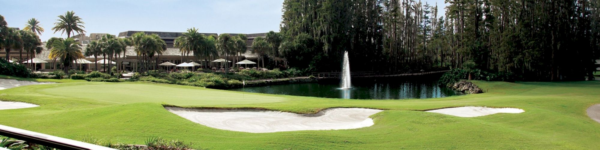 Lush green golf course with sand bunkers, a water fountain, surrounding trees, and buildings in the background under a clear sky.