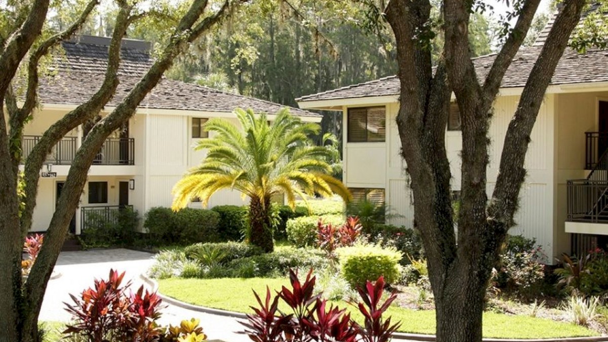 The image shows a peaceful garden area with trees, plants, and a bench, situated between two beige buildings under sunlight, ending the sentence.