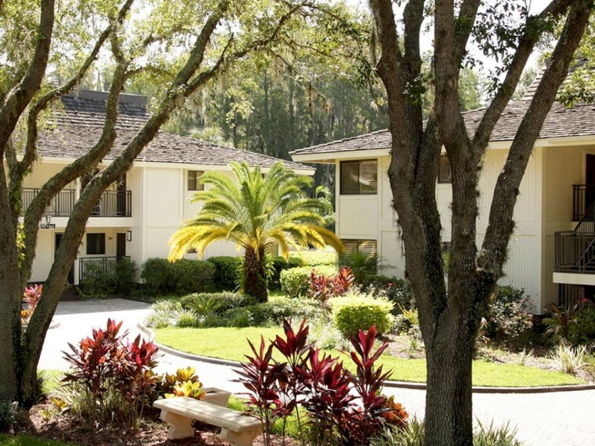 The image shows a peaceful garden area with trees, plants, and a bench, situated between two beige buildings under sunlight, ending the sentence.