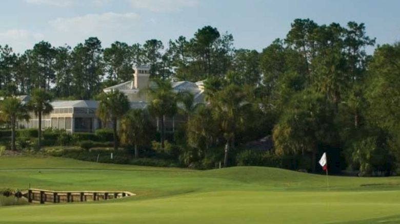 A scenic view of a golf course with a manicured green, surrounded by trees and water, and a clubhouse in the background.