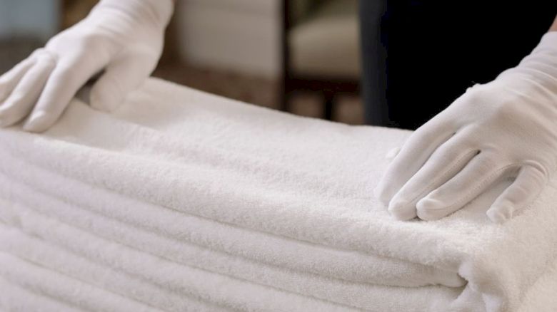 A person wearing white gloves is neatly stacking a pile of white towels on a table with a blue and gold patterned tablecloth.