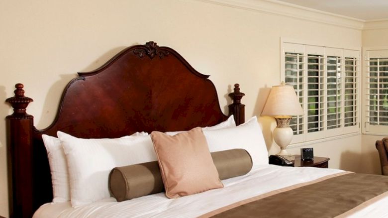 A cozy bedroom featuring a large bed with white linens, brown accents, a wooden headboard, nightstands, lamps, and window shutters.