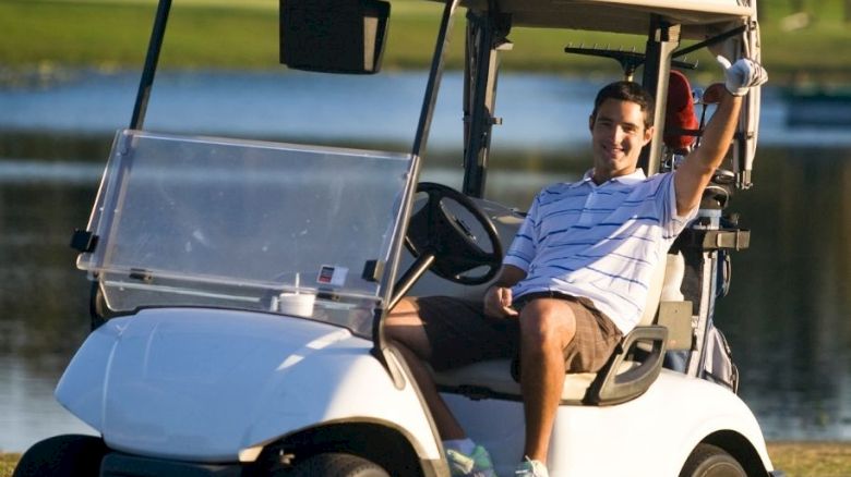 A person is sitting in a golf cart, raising their right hand, with a scenic view of a golf course and a lake in the background.