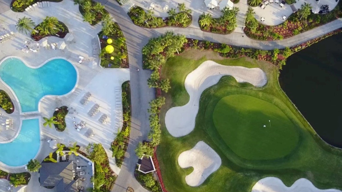 Aerial view of a resort with swimming pools, loungers, green landscaping, pathways, and a golf course with sand bunkers near a water body.