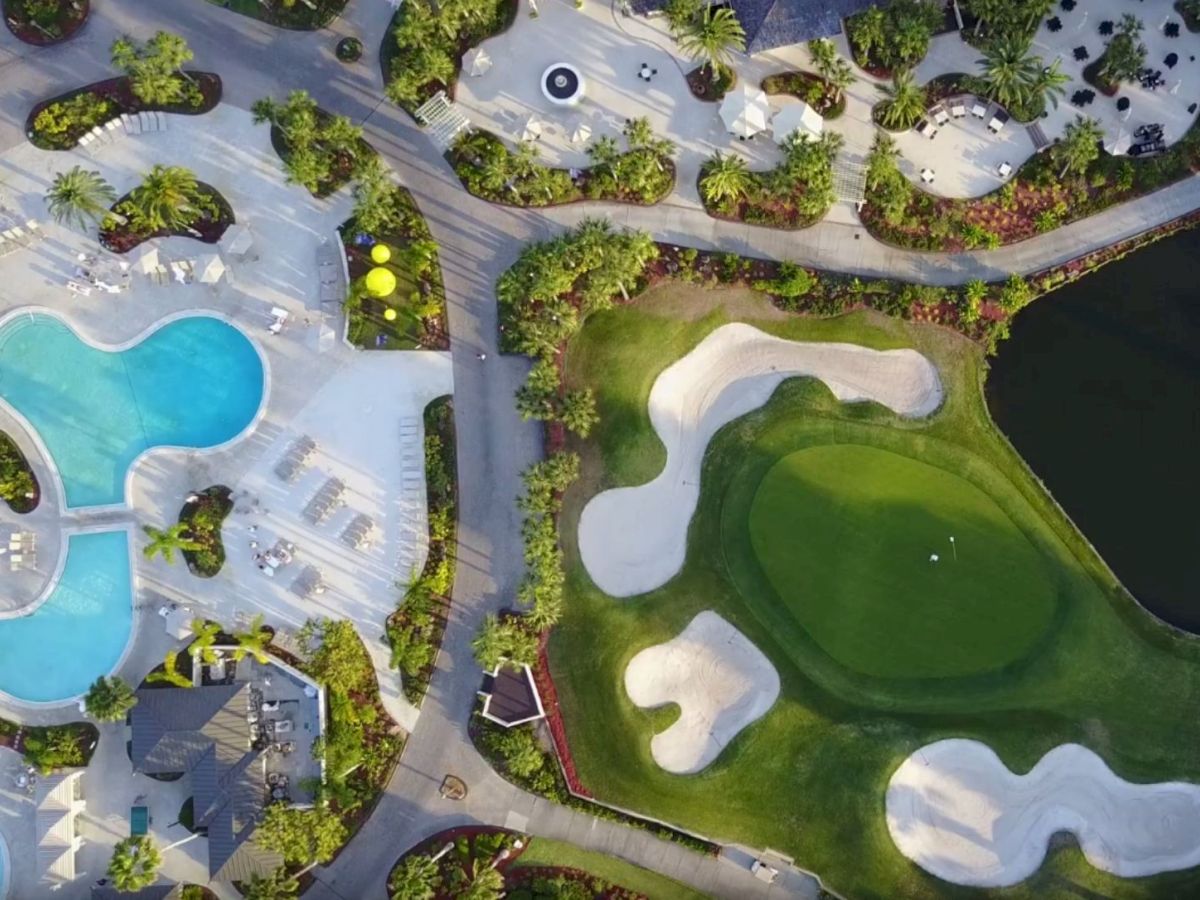 Aerial view of a resort with swimming pools, loungers, green landscaping, pathways, and a golf course with sand bunkers near a water body.