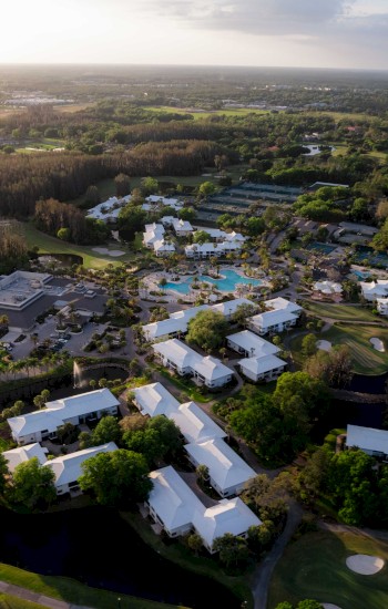 Aerial view of a resort with buildings and pools beside a golf course, surrounded by greenery.