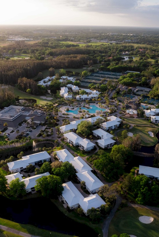 Aerial view of a resort with buildings and pools beside a golf course, surrounded by greenery.